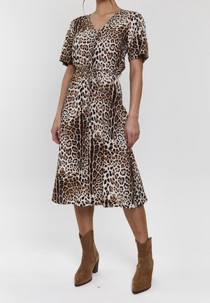Femme portant une robe imprimée léopard longueur genoux avec des manches courtes et des bottines en daim marron, debout devant un fond uni.