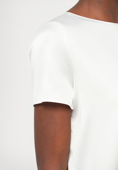 Λευκή μπλούζα κοντομάνικη με λε�ία υφή, στρογγυλή λαιμόκοψη και εφαρμοστή γραμμή, που τονίζει την περιοχή του ώμου και του άνω βραχίονα.