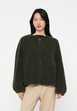 Frau in einer dunkelgrünen Fleecejacke mit Reißverschluss und gepufften Ärmeln, die beige Hosen trägt, steht vor einem schlichten weißen Hintergrund.