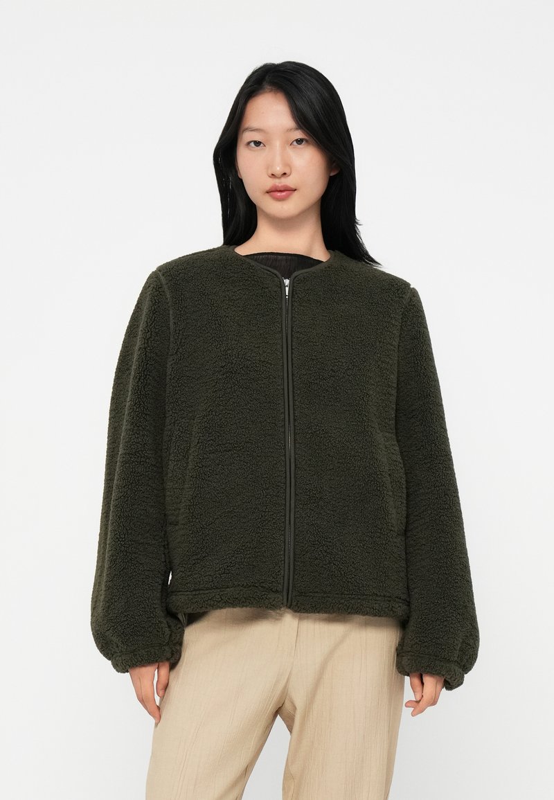 Woman wearing a dark green zip-up fleece jacket with puffed sleeves and beige pants, standing against a plain white background.