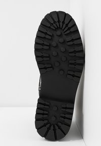 Semelle de botte noire avec des crampons profonds en forme hexagonale et angulaire conçus pour une forte adhérence, photographiée sur un fond blanc.