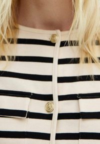 Gestreifter Cardigan in Beige und Schwarz mit rundem Ausschnitt, vorderem Knopfverschluss und zwei Brusttaschen mit goldfarbenen Knöpfen.