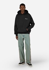 Felpa nera con cappuccio, tasca frontale e logo bianco, indossata con jeans a gamba larga azzurri chiari con bande laterali e sneaker nere.