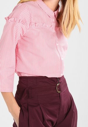 Femme portant un chemisier à rayures verticales roses et blanches avec un détail volanté et un pantalon taille haute bordeaux foncé avec une ceinture en tissu.