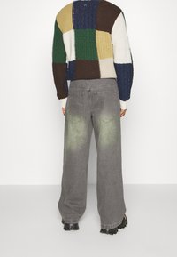 Suéter de gran tamaño y tejido tricotado con un diseño de patchwork multicolor, combinado con pantalones de mezclilla anchos grises que presentan un dobladillo verde desvaído y bolsillos traseros.