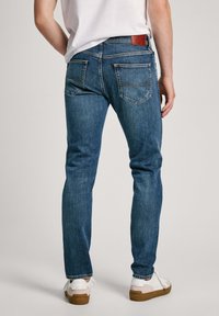 Jeans de mezclilla azul con corte ajustado, con bolsillos traseros y un parche de cuero con la marca. Llevados con zapatillas blancas sobre un fondo liso.