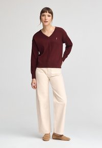 Burgunder v-ringad tröja med långa ärmar, parat med krämfärgade vida byxor. Tröjan har en liten logotyp på bröstet. Mocka slip-on skor.