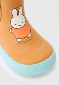 Botte en caoutchouc orange avec une semelle texturée bleu clair. Présente un autocollant de dessin animé d'un lapin en tenue orange sur le côté extérieur.