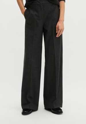 Woman wearing dark gray wide-leg trousers with side pockets and black shoes, standing against a plain light background.