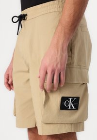 Beige Cargo-Shorts mit elastischem Bund und schwarzem Kordelzug. Verfügt über eine Seitentasche mit schwarzem Calvin Klein Logo-Patch. Strukturierter Stoff.