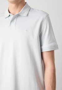 Homme portant un polo gris clair à manches courtes avec deux boutons et un petit logo "BOSS" brodé sur le côté gauche de la poitrine.