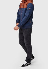 Homme portant une veste à capuche zippée bleu marine et rouille, un jean noir et des baskets noires et blanches, debout contre un fond clair uni.