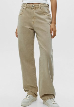 Personne portant un pantalon beige à jambes larges et des baskets blanches, debout devant un fond clair uni.