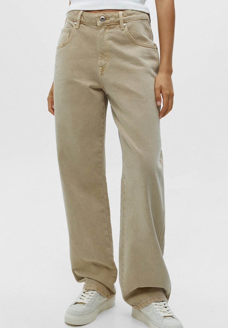 Personne portant un pantalon beige à jambes larges et des baskets blanches, debout devant un fond clair uni.