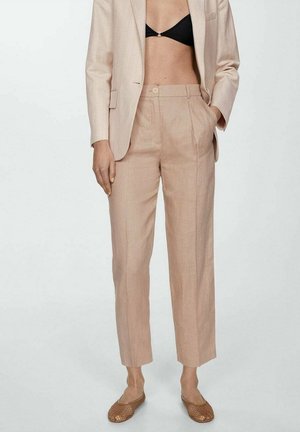 Vrouw draagt beige op maat gemaakte broek en bijpassend colbert met een zwarte bralette en bruine gevlochten ballerina's, staand tegen een effen achtergrond.