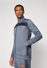 Grijze sportjack met zwarte accenten, voorzien van een hoge kraag, voorrits, twee zakken en oranje logo details op de borst en zoom.
