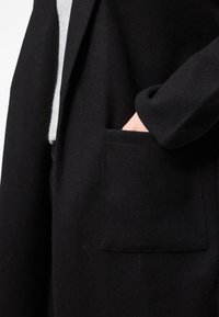 Abrigo de lana negro con un único bolsillo frontal, corte recto y mangas largas, con una textura suave y un diseño minimalista.