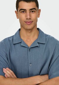 Korte mouwen, button-up shirt van gestructureerde blauwe stof met een kraag en een knoopdetails, gedragen door een mannelijke model met gekruiste armen.