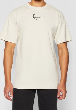 Beige puuvillainen t-paita, jossa on lyhyet hihat, pyöreä pääntie ja hillitty musta allekirjoituskuvio rintakehässä. Kevyt kangas, väljä leikkaus.