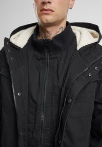 Chaqueta negra con un cierre frontal y solapa abotonada. Cuenta con una capucha y un cuello forrados de forro polar, tejido texturizado y múltiples bolsillos.