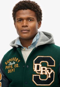 Groene en marineblauwe varsityjack met leren mouwen, voorzien van geborduurde tekst "SUPERDRY" en een grote "S" patch. Een grijze hoodie steekt uit de kraag.