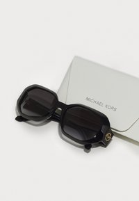 Juodi Michael Kors akiniai nuo saulės su aštuonkampiu rėmu, tamsiais lęšiais ir auksiniu logotipo akcentu. Komplekte yra baltas tekstūruotas dėklas.