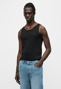 Μαύρο βαμβακερό τοπ χωρίς μανίκια με φαρδύ λαιμό και λεπτή εφαρμογή, συνδυασμένο με ανοιχτό μπλε τζιν παντελόνι. Διακριτικό μικρό λογότυπο στο στήθος.