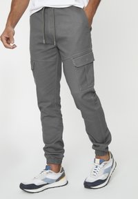 Grijze cargobroek met een elastische tailleband, taps toelopende boorden en meerdere zijzakken, gecombineerd met witte en marineblauwe sportschoenen.