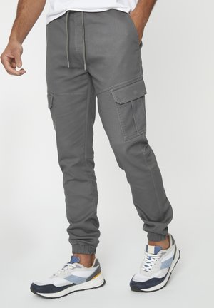 Grijze cargobroek met een elastische tailleband, taps toelopende boorden en meerdere zijzakken, gecombineerd met witte en marineblauwe sportschoenen.