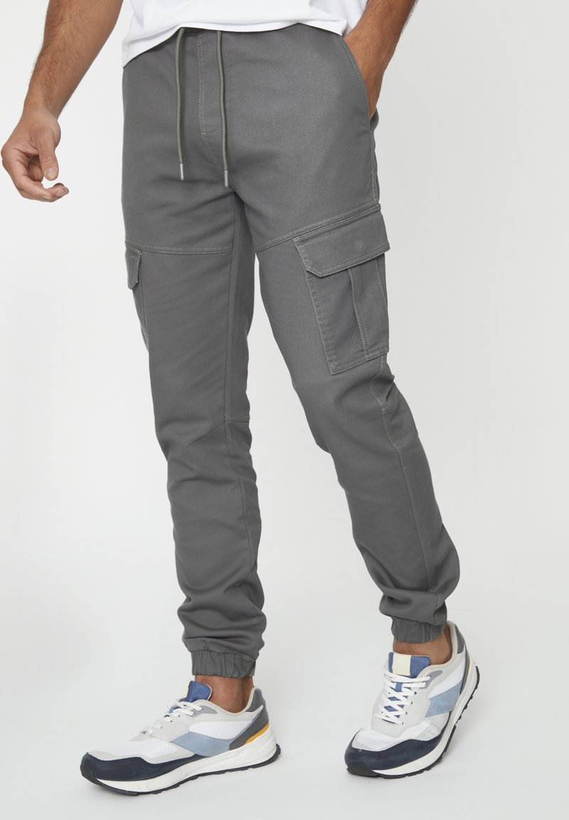 Grijze cargobroek met een elastische tailleband, taps toelopende boorden en meerdere zijzakken, gecombineerd met witte en marineblauwe sportschoenen.