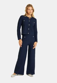 Ensemble bleu marine assorti comprenant une veste courte texturée avec quatre poches avant et des boutons métalliques, associé à un pantalon large.