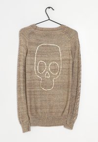 Beige stickad tröja med en texturerad dödskalle-design i metalltråd på baksidan. Inkluderar ribbade muddar och halsringning.