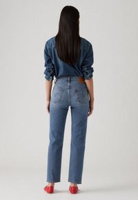 Pantalones vaqueros de mezclilla en azul claro, con cintura alta, corte recto y bolsillos traseros. Combinados con una camisa de mezclilla azul oscuro. Zapatos rojos.