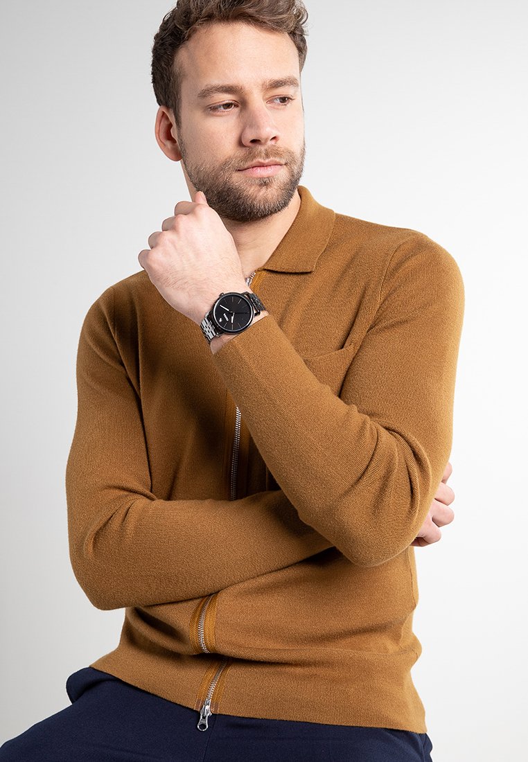 Suéter marrón de cremallera hecho de un suave material de punto con cuello, que cuenta con un bolsillo delantero y mangas largas, usado sobre pantalones azul marino.