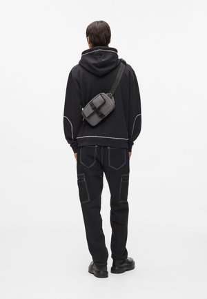 Sudadera con capucha negra y costuras blancas, combinada con pantalones cargo negros y una bolsa cruzada gris con hebilla y correa ajustable.