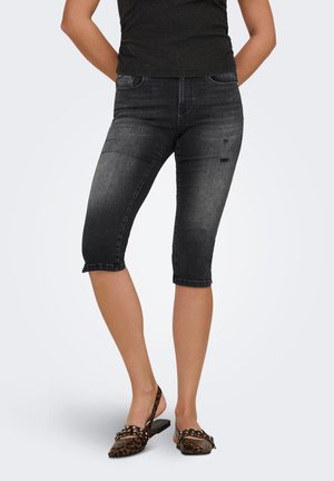Vrouw die zwarte cropped skinny jeans draagt met lichte slijtage en platte slingbacks met luipaardprint, staand met handen achter de rug.