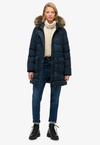 Giacca puffer blu navy con cappuccio foderato in pelliccia ecologica, chiusura frontale a scatto, due ampie tasche e maglione in maglia testurizzata sotto.