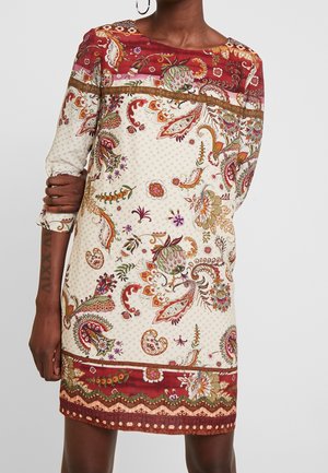 Mujer con vestido crema con patrones florales de cachemira en rojo, naranja y verde y mangas tres cuartos, de pie con un brazo cruzado.