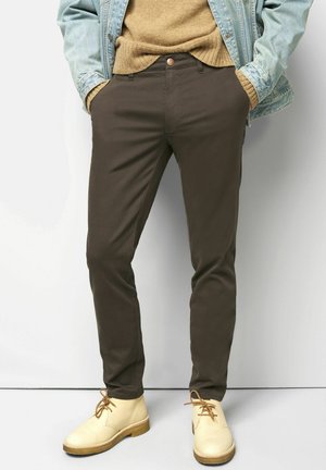 Braune Baumwollhose mit schmalem Schnitt, mit Knopfverschluss, Seitentaschen und kombiniert mit hellbeigen Schuhen.