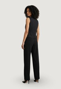 Femme aux cheveux bouclés portant un haut noir sans manches, un pantalon large noir et des talons hauts noirs, debout et regardant par-dessus son épaule.