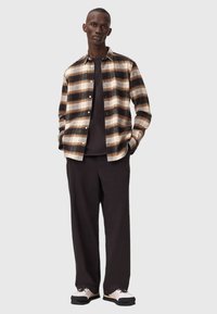 Camicia a scacchi in marrone e nero, abbinata a una maglietta nera e pantaloni larghi neri. Le scarpe presentano un design bianco e nero.