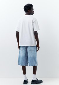 Jeune homme aux cheveux courts et bouclés, portant un t-shirt blanc ample, un short en jean long, des chaussettes blanches et des mocassins noirs, debout contre un mur uni.