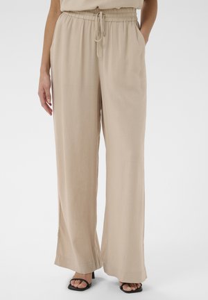 Pantalon beige à jambes larges avec taille élastique et cordon de serrage, porté avec des sandales noires à talons et orteils découverts, mains dans les poches.