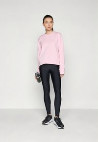 Sudadera rosa claro con puños acanalados, combinada con leggings negros y zapatillas deportivas negras. Sosteniendo una botella de agua gris.