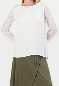 Femme portant une blouse blanche à manches longues transparentes et à motifs, ainsi qu'une jupe portefeuille verte olive avec un nœud sur le côté.