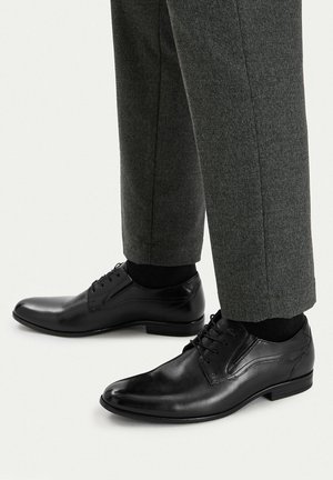 Czarne skórzane eleganckie buty noszone z ciemnoszarymi formalnymi spodniami i czarnymi skarpetkami na jasnym tle.