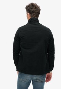 Zwarte katoenen jas met een hoge kraag en lange mouwen, met een eenvoudige textuur en een licht gebogen zoom. Draagt over een blauwe spijkerbroek.