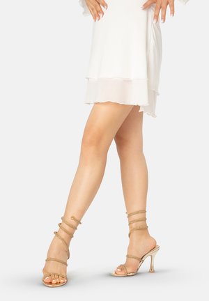 Piernas con sandalias doradas de tacón alto con tiras combinadas con una minifalda blanca de capas, manos apoyadas en las caderas sobre un fondo claro.