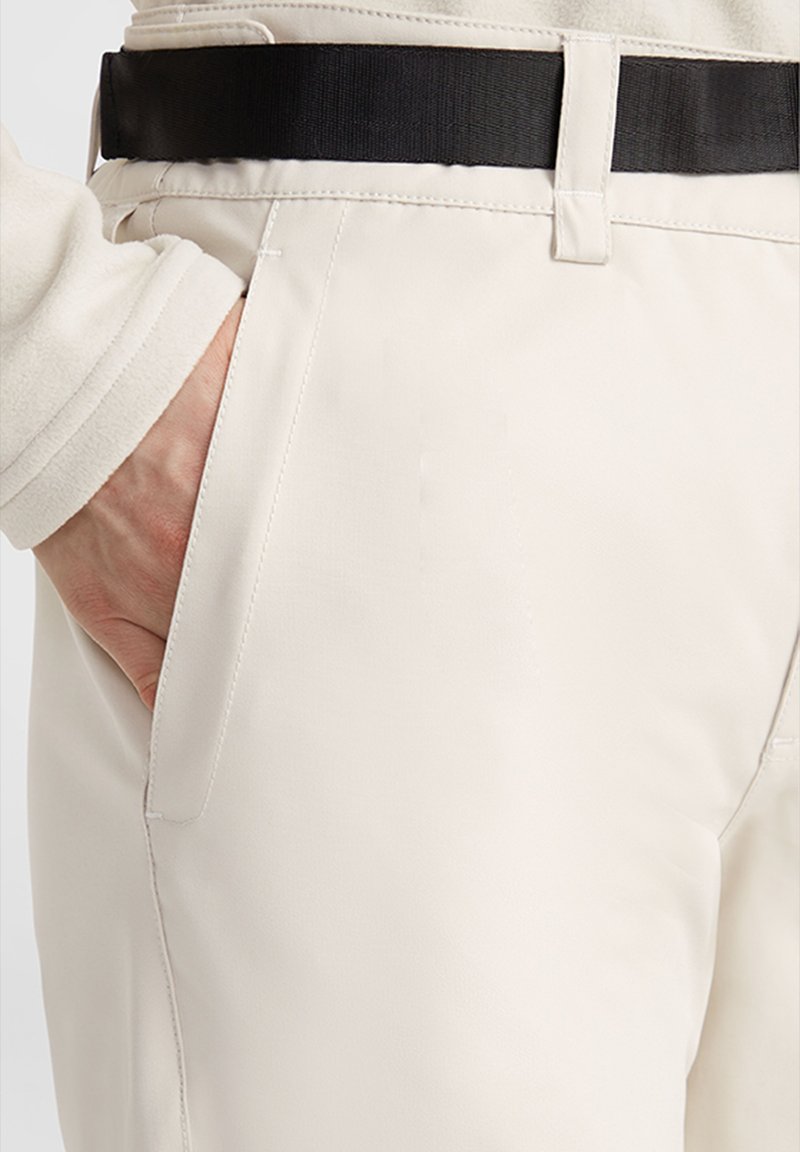 Pantalones beige claros con una textura suave, que cuentan con un cinturón negro y bolsillos laterales, mostrados del cinturón hacia abajo con la mano en el bolsillo.
