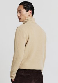 Uomo con capelli lisci all'indietro, che indossa un maglione a collo alto beige e pantaloni marrone scuro, che guarda di lato su uno sfondo semplice.